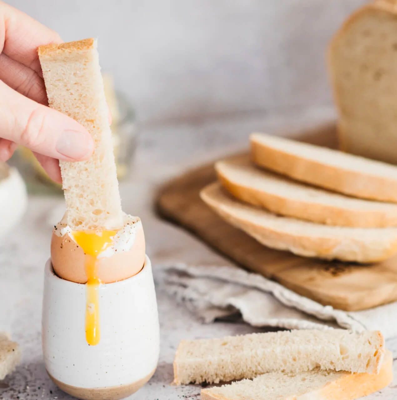 A freshly baked white loaf of bread cut into slices sits in the background. In the foreground, soldiers are dipped into a boiled egg.