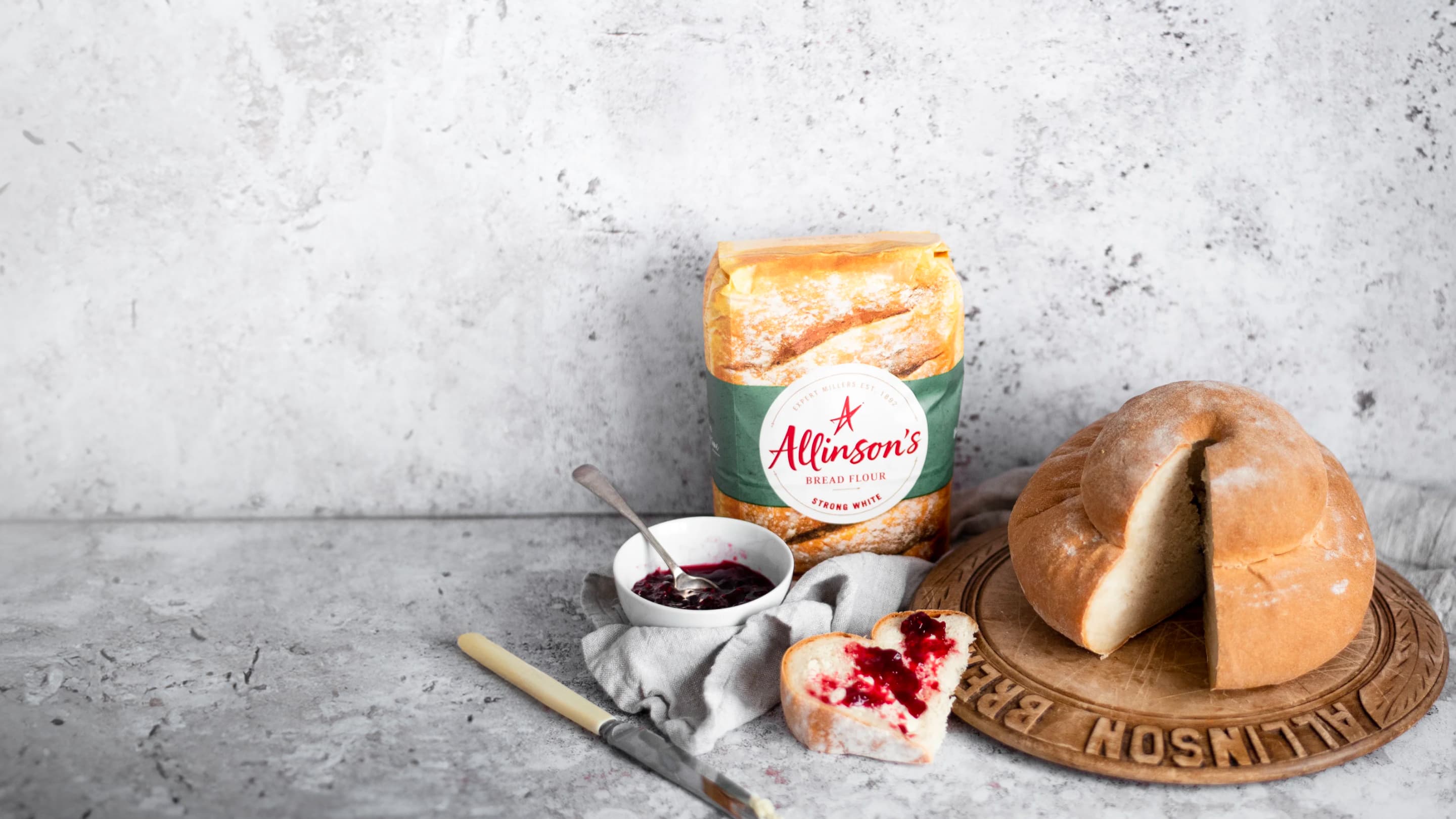 A freshly baked cottage loaf on a bread board site alongside a pack of Allinson's Strong White Bread Flour. A slice had been cut and spread with butter and jam.